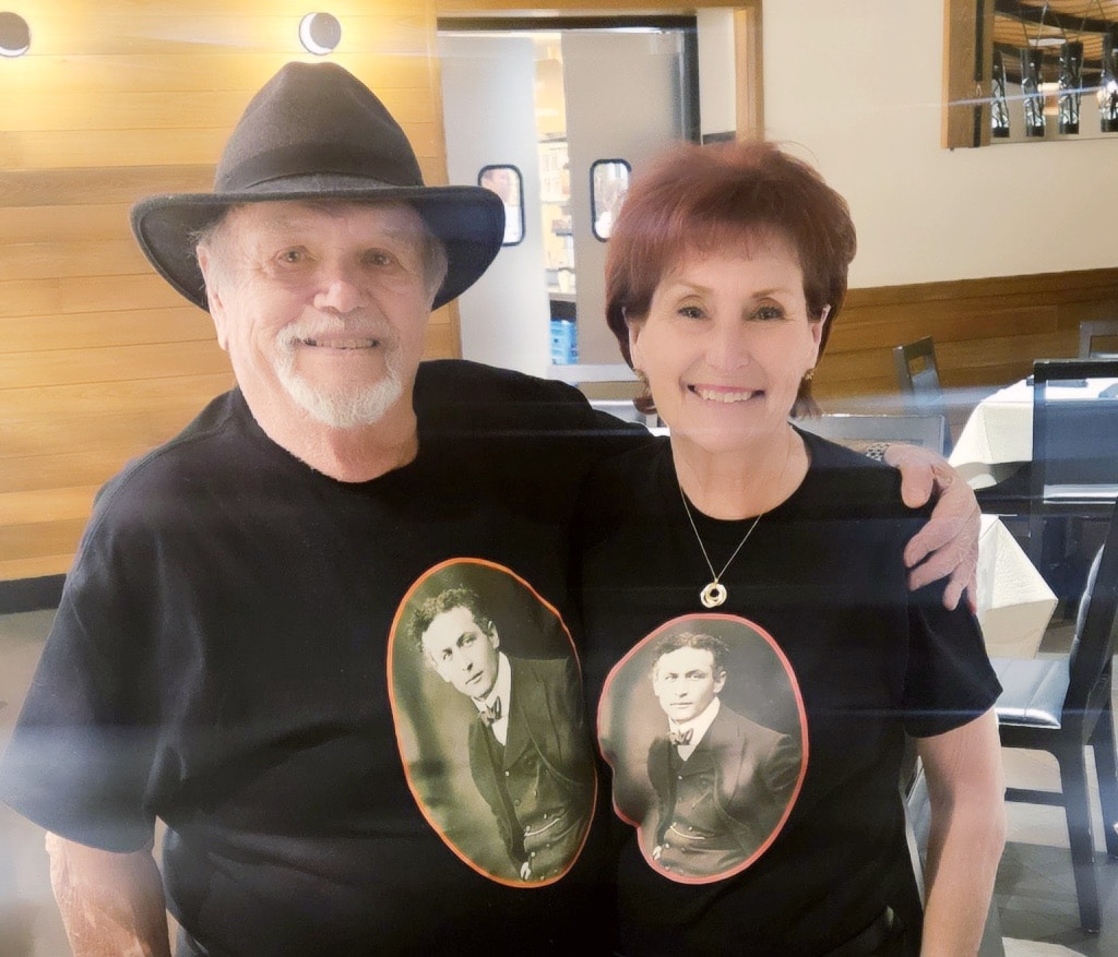 Older couple wearing black t-shirts with vintage portraits; man wears a black hat, both smiling, in a restaurant setting.