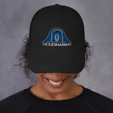 Person wearing a black cap with "Houdinarian" and a design. Face partially visible, looking down, gray background.