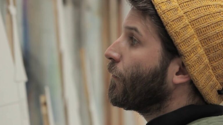 Profile of a person with a beard wearing a yellow knit beanie, looking intently at something out of frame indoors.