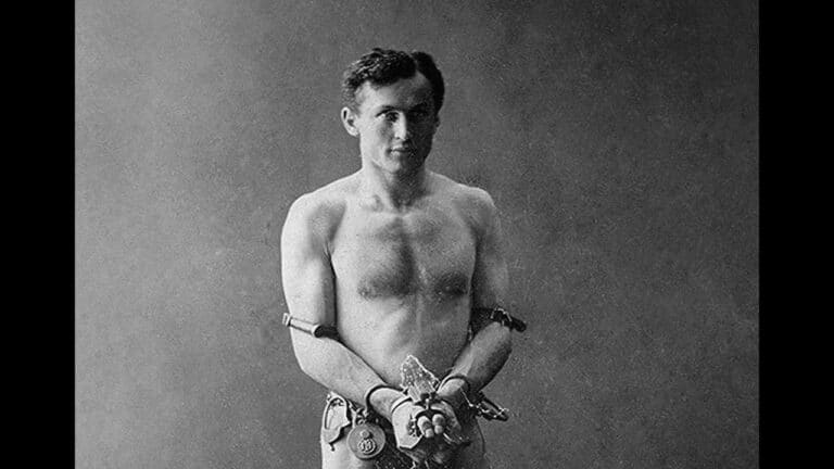 A shirtless man stands with his hands and arms restrained by chains and locks.