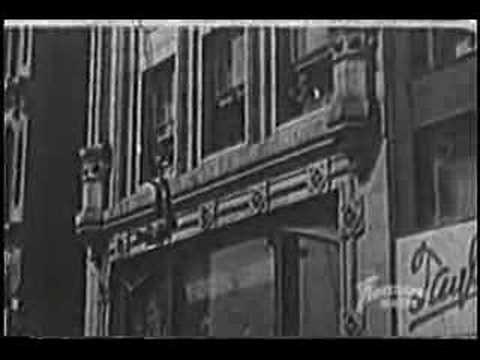 Black and white photo of a man hanging from a building ledge, with ornate architectural details and a sign partially visible.