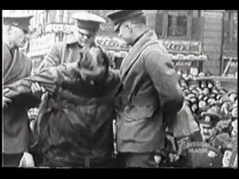 Historic black and white photo of a crowd watching military personnel helping a person in a jacket, possibly in a public space.