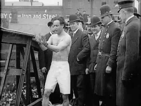 A man in shorts performs a stunt on a bridge, surrounded by onlookers and police officers in early 20th-century attire.