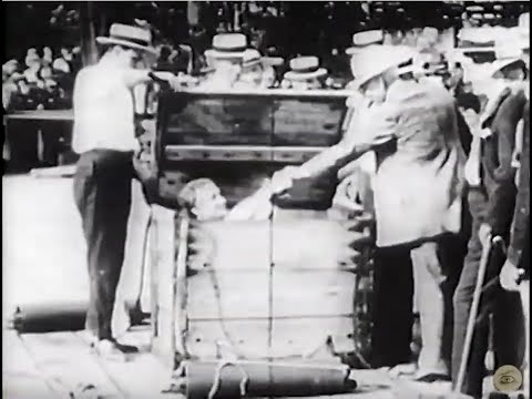 A person is submerged in a box filled with water, while being assisted by two individuals, surrounded by a crowd.