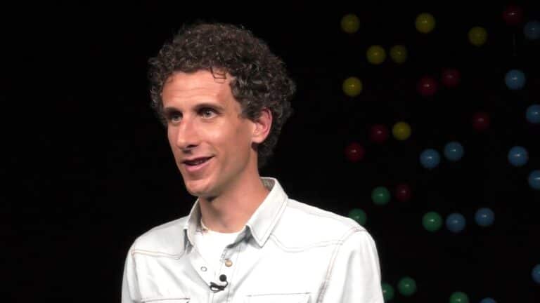 Person with curly hair and a microphone, wearing a light-colored shirt, speaks against a dark background with colorful dots.