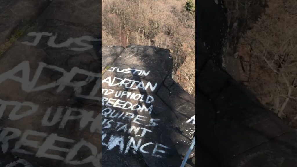 Graffiti on a rock with phrases: "Justin Adrian to uphold freedom" in white paint, surrounded by trees.