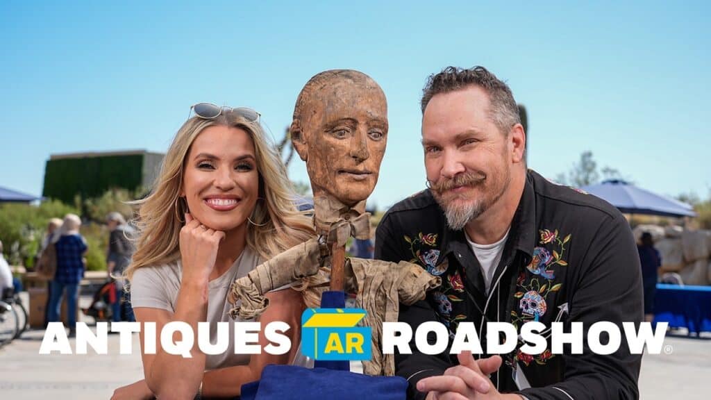 Two people pose with a wooden puppet between them. The "Antiques Roadshow" logo is at the bottom.