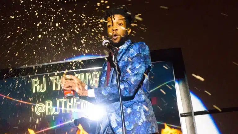 A man in a blue patterned suit reacts as liquid sprays from a can he's holding, standing in front of a screen with text.