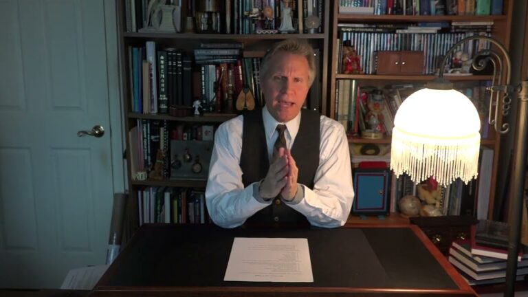 Person seated at a desk with hands clasped, against a backdrop of a filled bookshelf and a lit table lamp, facing the camera.