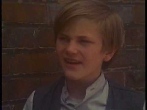 Young person with short hair wearing a vest and shirt, standing in front of a brick wall.