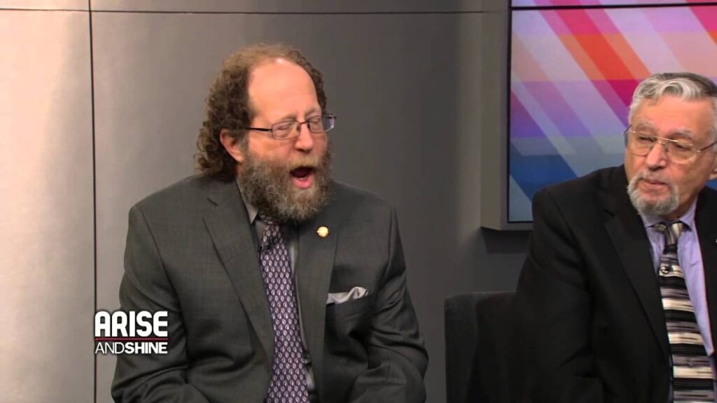 Two men in suits sit together on a talk show set, one with a beard and glasses is speaking, while the older man with glasses listens. The screen in the background has a colorful geometric design.