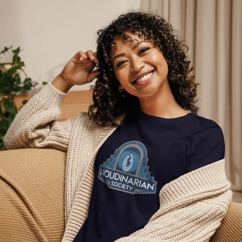 Smiling woman with curly hair wears a navy "Houdinarian Society" T-shirt and a knitted cardigan, sitting in a room with plants and curtains.