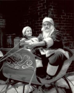 Santa Claus and a child in Santa hats sit in a decorated sleigh, holding reins, with a brick wall in the background.