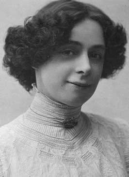 A woman from the early 20th century with curly hair, wearing a high-necked lace blouse, faces the camera with a slight smile.