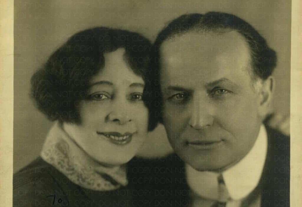 A vintage sepia-toned portrait of a smiling woman with curly hair and a stern-looking man, both wearing early 20th-century attire, closely positioned side by side.