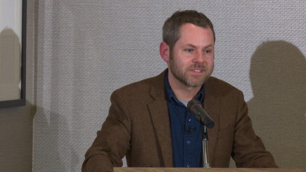 Man with a beard speaking at a podium with a microphone, wearing a brown jacket and blue shirt. Textured wall in the background.