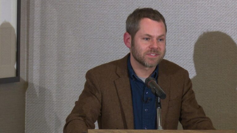 Man with a beard speaking at a podium with a microphone, wearing a brown jacket and blue shirt. Textured wall in the background.