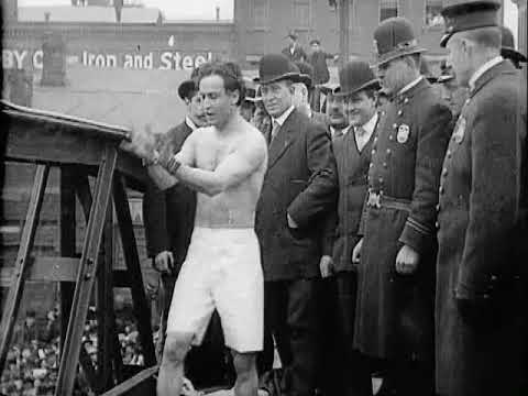 A man in shorts and chains prepares to perform a stunt in front of a group of onlookers and policemen in early 20th-century attire.