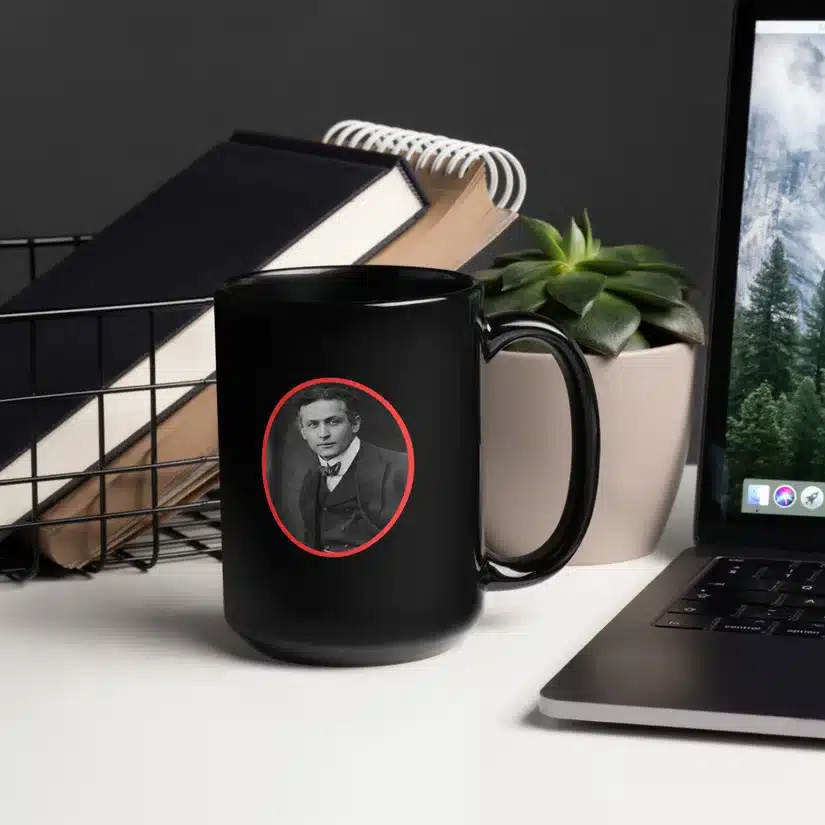 Black mug with a portrait of a man in a suit, placed on a desk next to books, a succulent plant, and a laptop.