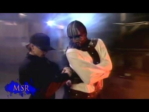 A person in a black outfit is fastening a straitjacket on another individual wearing face paint and the straitjacket.