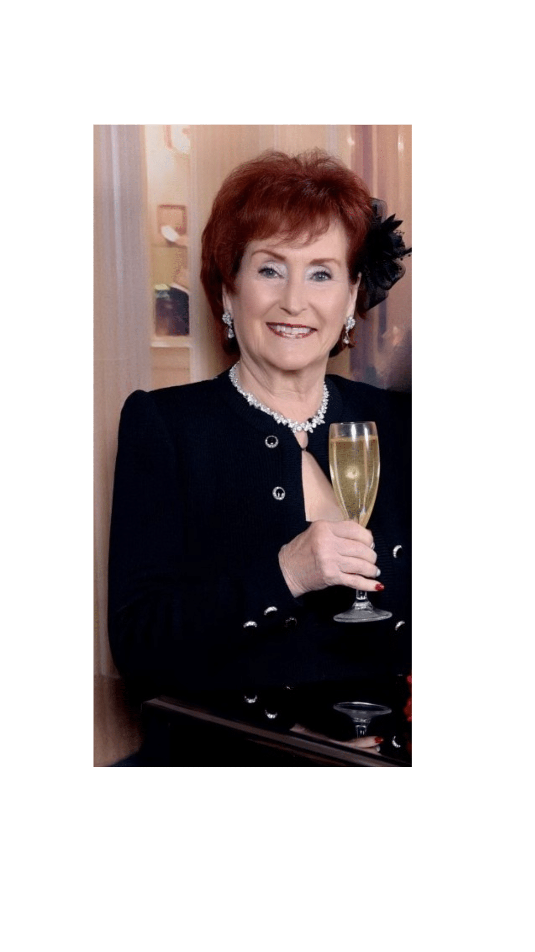 A woman with short red hair is smiling and holding a glass of champagne, wearing a black outfit and pearl necklace.