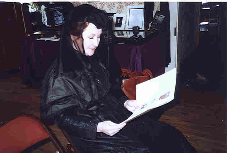 A person in dark clothing and a hat with a veil, seated in a chair, is reading a large document or book. The background includes a table and framed photographs.