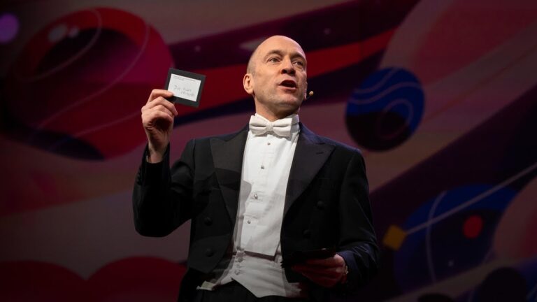 A bald man in a tuxedo holds up a small card on stage. Colorful abstract background.