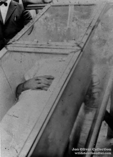 A person lies inside a glass box with their hands folded on their chest. The box is transparent, revealing a white garment. The image is black and white.