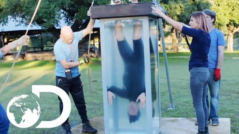 Person upside down in a transparent water tank outdoors, surrounded by four people assisting.