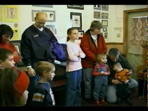 A group of adults and children stand together indoors near a wall with framed pictures and a door with patterned curtains.