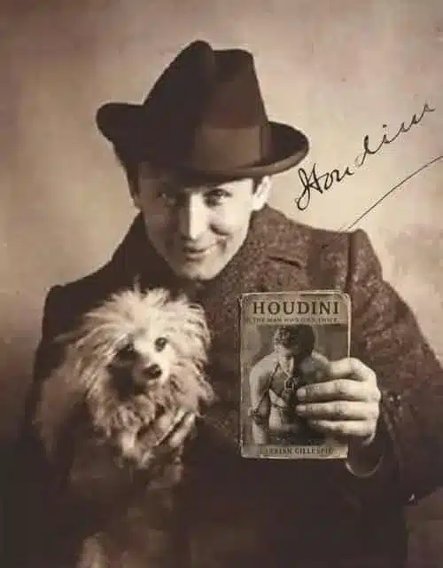 A man in a hat holds a small, fluffy dog and a book titled "Houdini: The Man Who Walked Through Walls" by Gibson. A signature is written on the image.