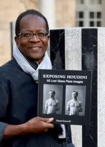 A man wearing glasses and a scarf holds a book titled "Exposing Houdini: 52 Lost Glass Plate Images," featuring two black-and-white images of Houdini on the cover.