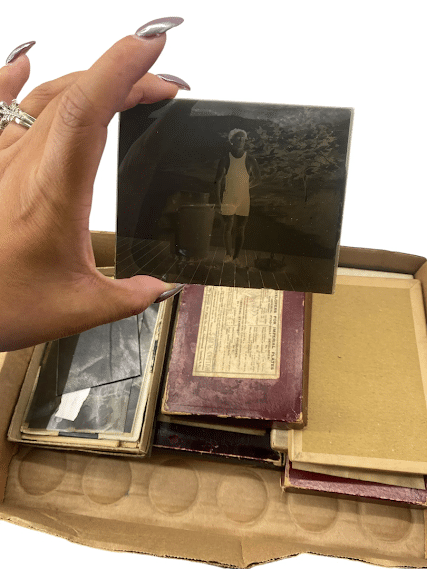 A hand holds up a black-and-white photo negative above a box filled with old photographic plates and documents.